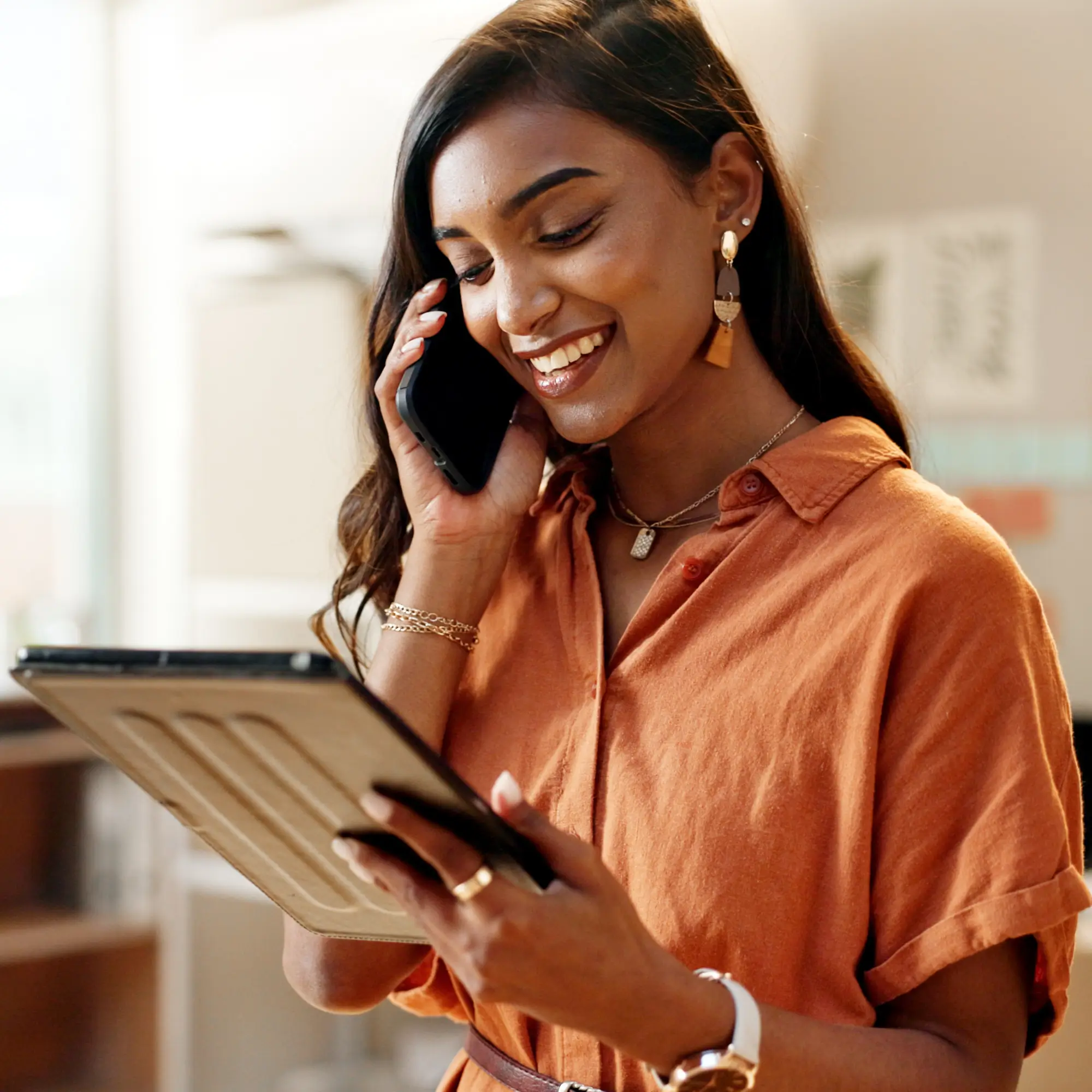 A smiling woman wearing a rust-colored dress, stylish earrings, and layered necklaces, standing indoors while talking on a smartphone and holding a digital tablet, appearing engaged with an AI voice agent to manage her tasks efficiently.