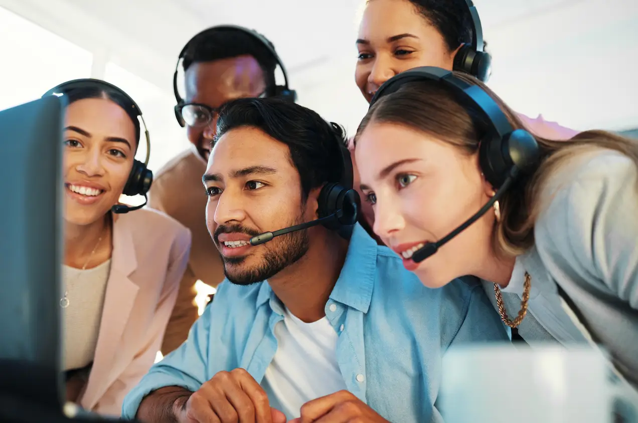 Group of customer service agents wearing headsets smiling and collaborating around a computer, showing teamwork in knowledge management for customer service.